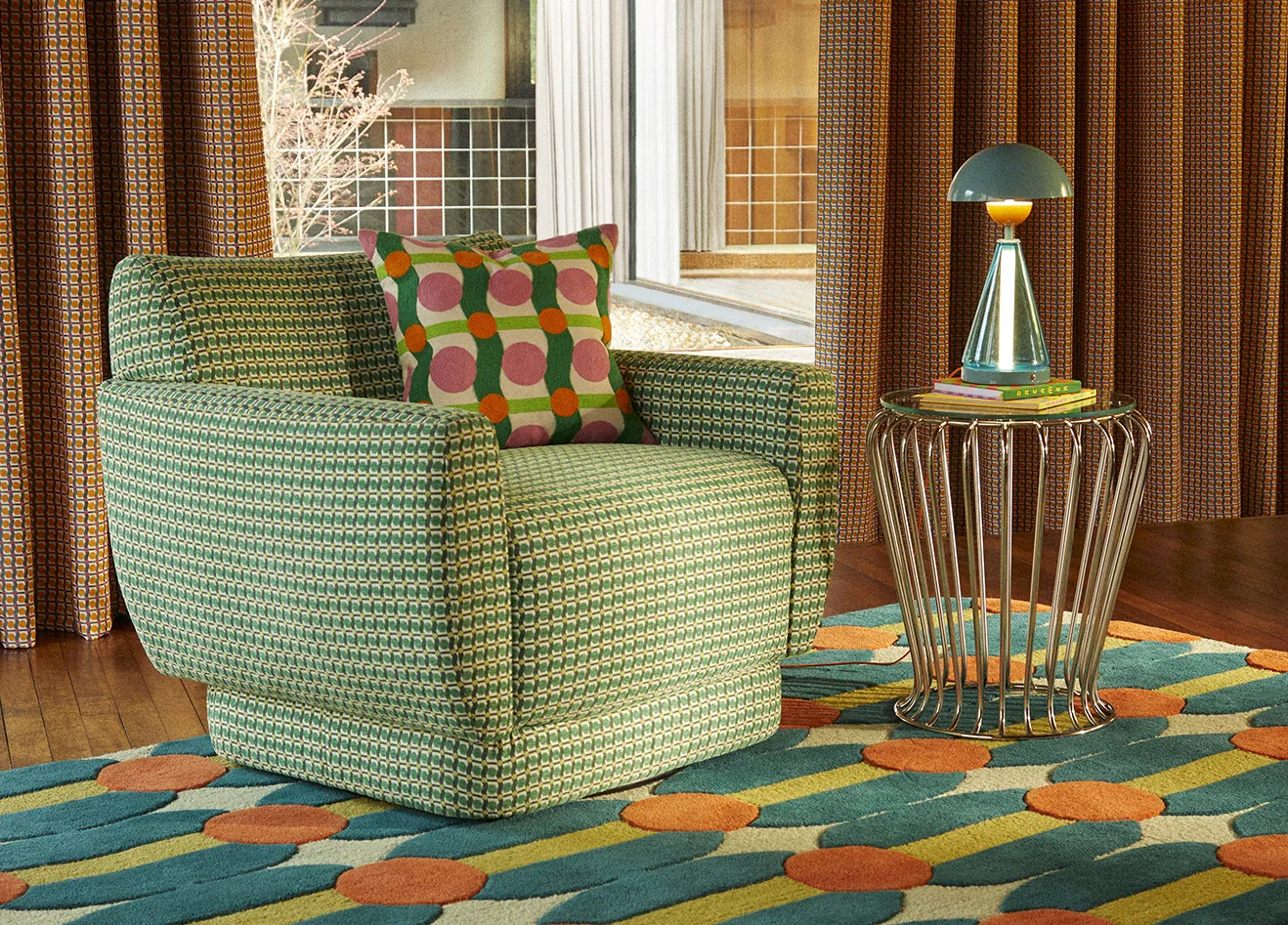 Seating area with a green patterned armchair, a colorful geometric area rug, patterned curtains, and a cone-base table lamp on a wire table.